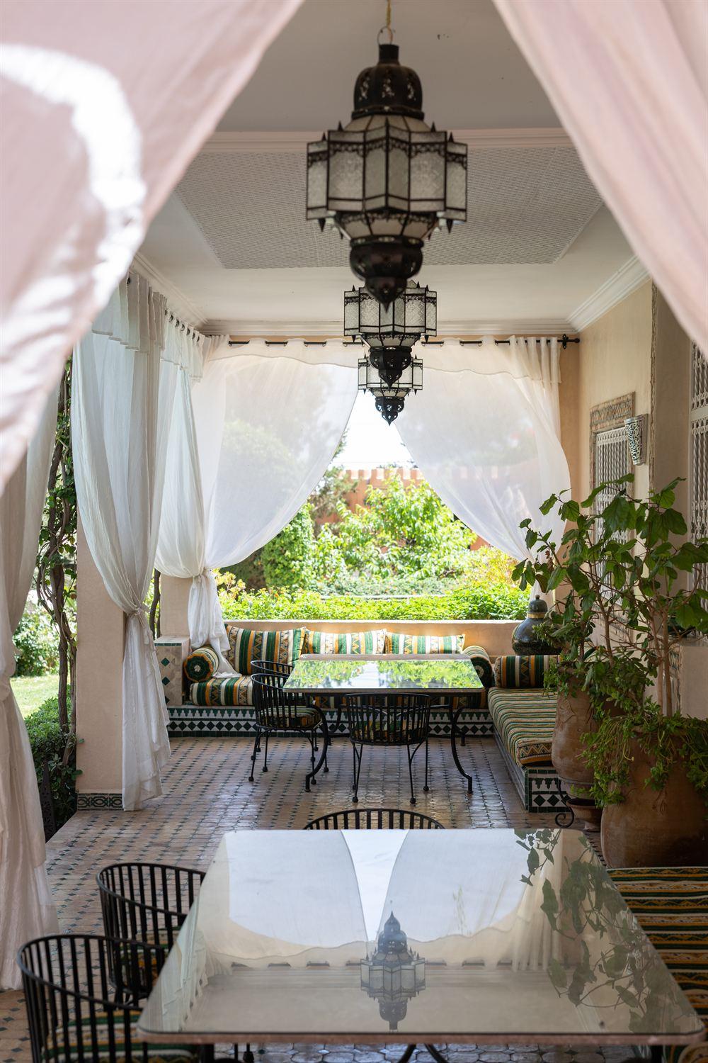 La terrasse d'un établissement : voilages blancs, lanternes, banquettes en zellige, jardin en arrière-plan.